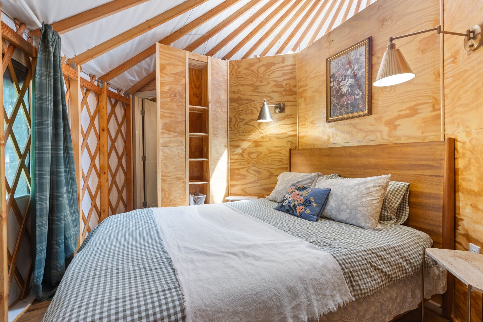 A cozy bed with a checker-pattern blanket in a luxurious circular yurt on a farm in Ellijay, Georgia. The space features two elegant golden lamps, a framed floral picture, and warm ambient lighting, offering a unique and comfortable stay for overnight rentals.