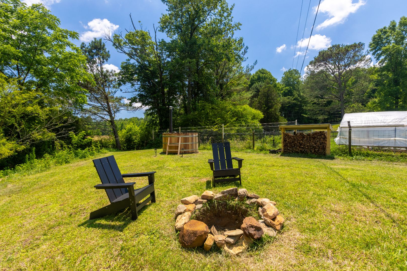 Relaxing evening at Bereishis The Farm in Ellijay, GA: A cozy bonfire surrounded by two black lounge chairs, with a wood-fired sauna nearby. The serene setting includes a lush garden and creek, offering a peaceful retreat at this fully organic farm that hosts farm stays, tours, and photoshoots.