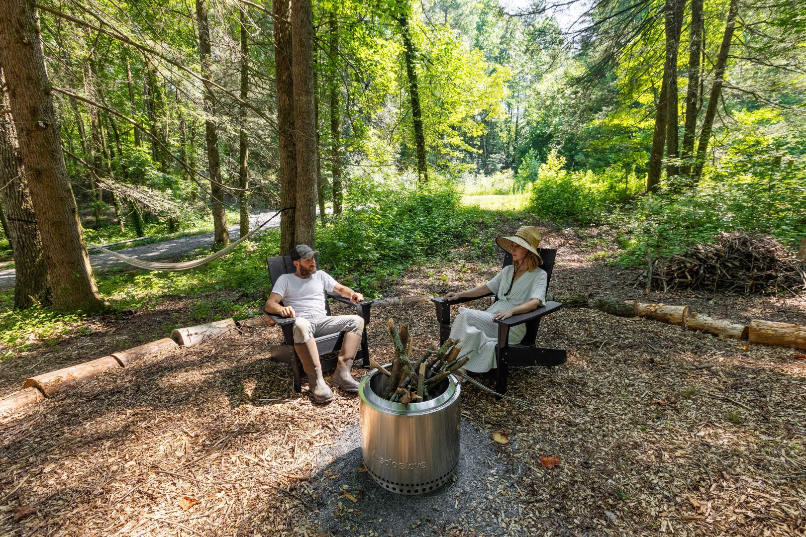 Outdoor scenic bonfire area with two black lounge chairs in front of a luxury yurt on Bereishis The Farm in Ellijay, Georgia. A farm couple relaxes in the chairs, enjoying the peaceful setting. The yurt is available for rent for a night or more, offering a serene escape on the organic farm.